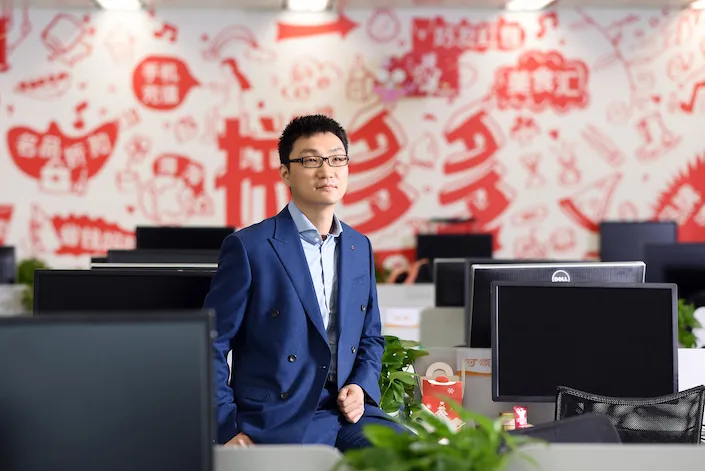 Man in a blue suit perched on a desk in an open office, in front of a red-and-white mural of Chinese shopping slogans