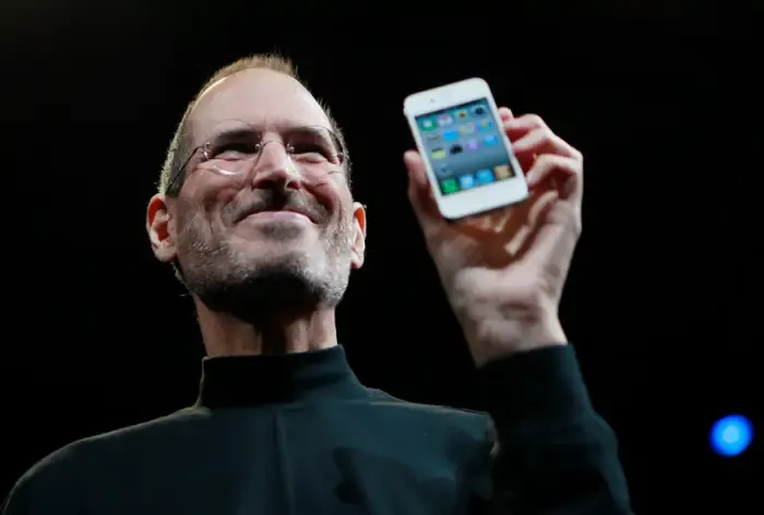 Presenter on a dark stage smiling as he holds up a white iPhone 4