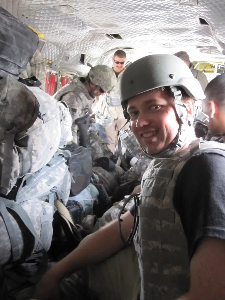 Smiling man in a helmet and body armor over a T-shirt, squeezed among uniformed troops and piled gear inside a military aircraft