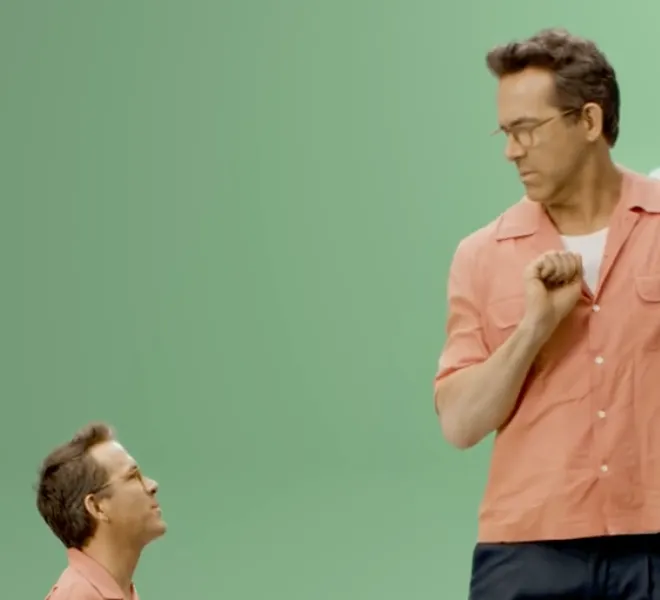 Ryan Reynolds shown twice against a green screen, a small kneeling version looking up at a full-size version in a coral shirt